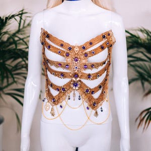 May include: A gold-colored, beaded and jeweled corset with purple accents. The corset is on a white mannequin.