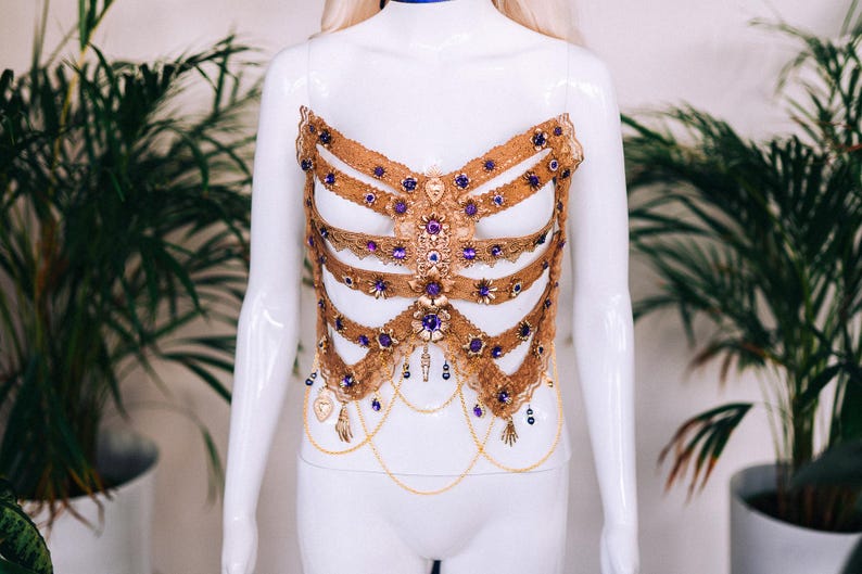 May include: A gold-colored, beaded and jeweled corset with purple accents. The corset is on a white mannequin.