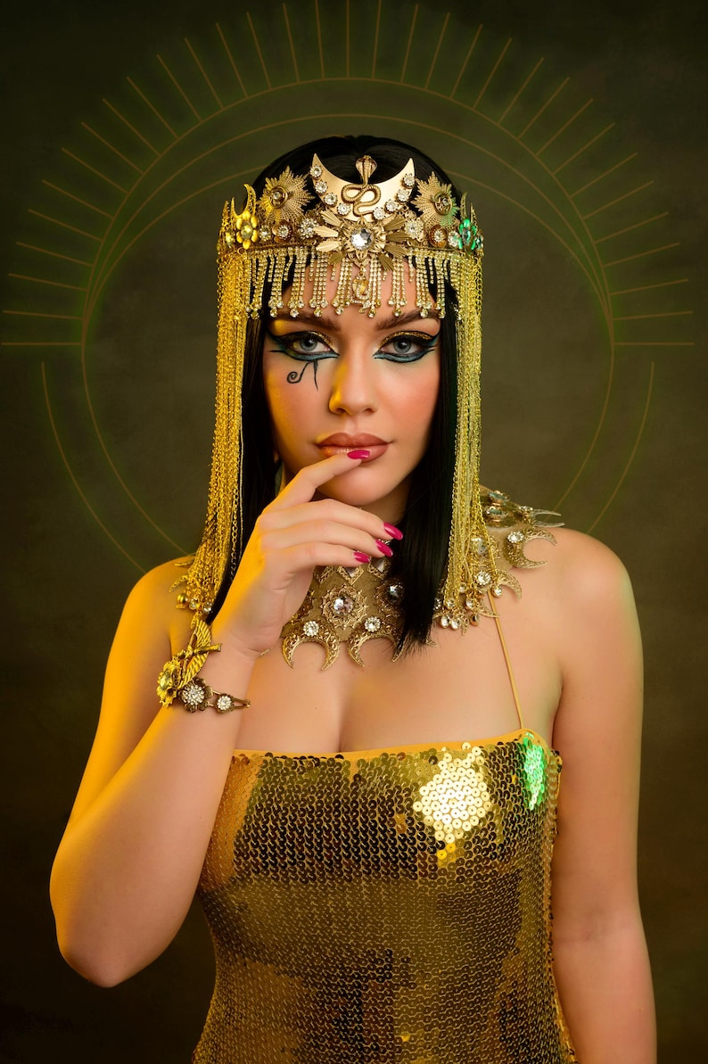 Peut inclure: Une femme portant une robe paillet&eacute;e dor&eacute;e et une couronne dor&eacute;e avec un motif de serpent. Elle a les cheveux noirs et un maquillage des yeux fonc&eacute;.