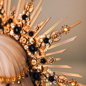 May include: A gold-colored crown with intricate detailing, featuring dark blue gemstones and clear crystals. The crown has a sunburst design with pointed rays and floral accents. A chain with dangling beads is also visible.