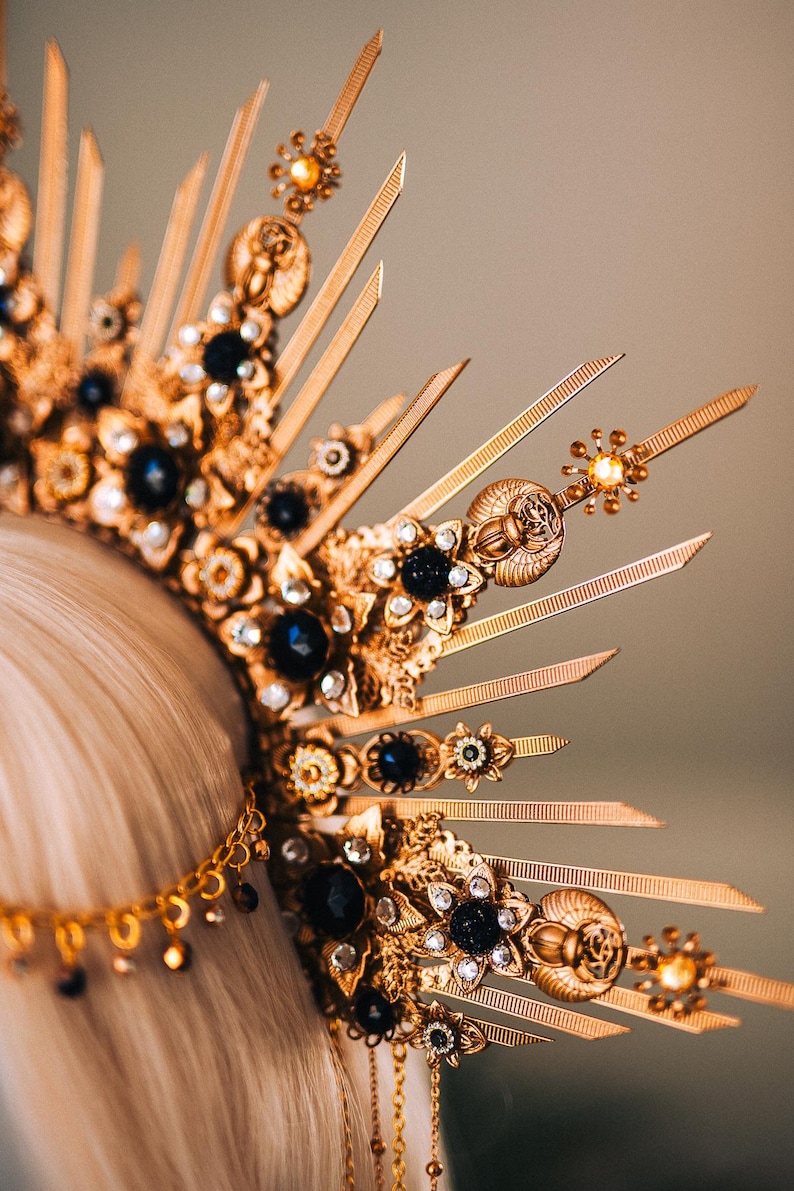 May include: A gold-colored crown with intricate detailing, featuring dark blue gemstones and clear crystals. The crown has a sunburst design with pointed rays and floral accents. A chain with dangling beads is also visible.