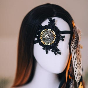 Puede incluir: M&aacute;scara de ojo de encaje negro con un dise&ntilde;o circular y calavera dorada. La m&aacute;scara est&aacute; adornada con encaje negro y una banda el&aacute;stica negra. Plumas en tonos marr&oacute;n, negro y blanco est&aacute;n unidas al lado.