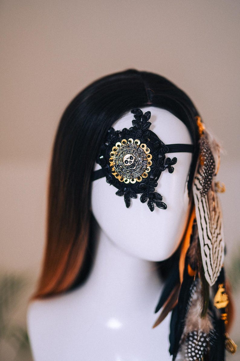 Puede incluir: M&aacute;scara de ojo de encaje negro con un dise&ntilde;o circular y calavera dorada. La m&aacute;scara est&aacute; adornada con encaje negro y una banda el&aacute;stica negra. Plumas en tonos marr&oacute;n, negro y blanco est&aacute;n unidas al lado.