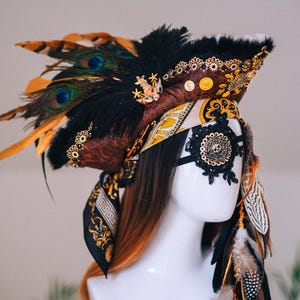 Puede incluir: Tocado decorativo con tem&aacute;tica pirata. El tocado presenta un sombrero marr&oacute;n y negro adornado con plumas de pavo real, adornos dorados y una m&aacute;scara para los ojos de encaje negro con un dise&ntilde;o de calavera. Una bufanda estampada est&aacute; envuelta alrededor de la cabeza.