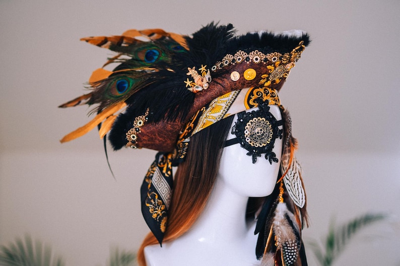 Puede incluir: Tocado decorativo con tem&aacute;tica pirata. El tocado presenta un sombrero marr&oacute;n y negro adornado con plumas de pavo real, adornos dorados y una m&aacute;scara para los ojos de encaje negro con un dise&ntilde;o de calavera. Una bufanda estampada est&aacute; envuelta alrededor de la cabeza.
