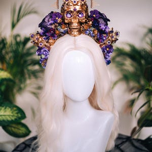 May include: A gold skull crown with purple flowers and jewels. The crown is on a white mannequin head with blonde hair.