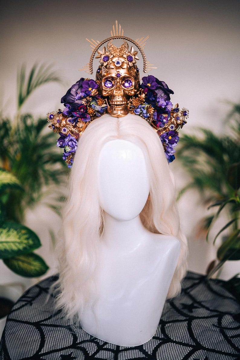 May include: A gold skull crown with purple flowers and jewels. The crown is on a white mannequin head with blonde hair.