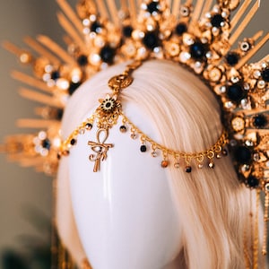 May include: A gold-colored headpiece with black and clear gemstones, and an Ankh pendant. The headpiece is on a mannequin head with blonde hair. The headpiece has a sunburst design.