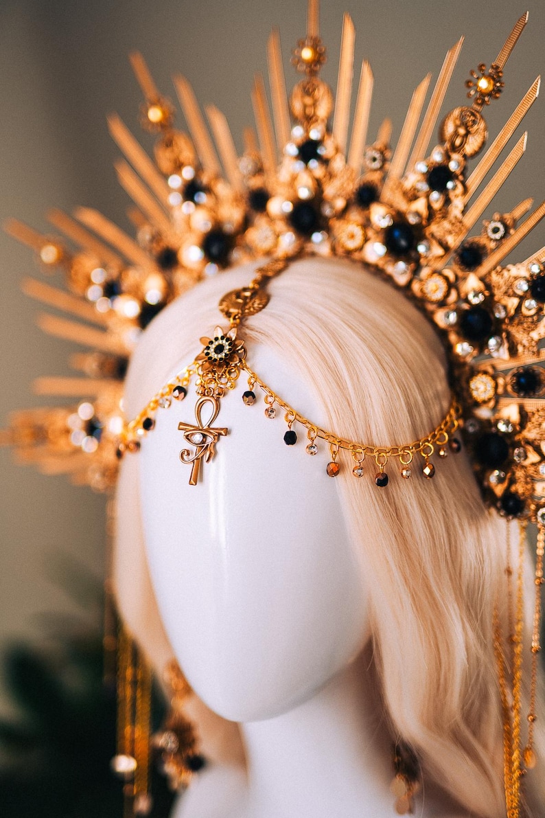 May include: A gold-colored headpiece with black and clear gemstones, and an Ankh pendant. The headpiece is on a mannequin head with blonde hair. The headpiece has a sunburst design.