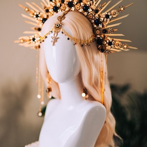 May include: A gold-toned headpiece with a sunburst design, adorned with dark blue gemstones and dangling chains. The headpiece features an Ankh symbol and is displayed on a white mannequin head with blonde hair, showcasing a celestial or fantasy theme.