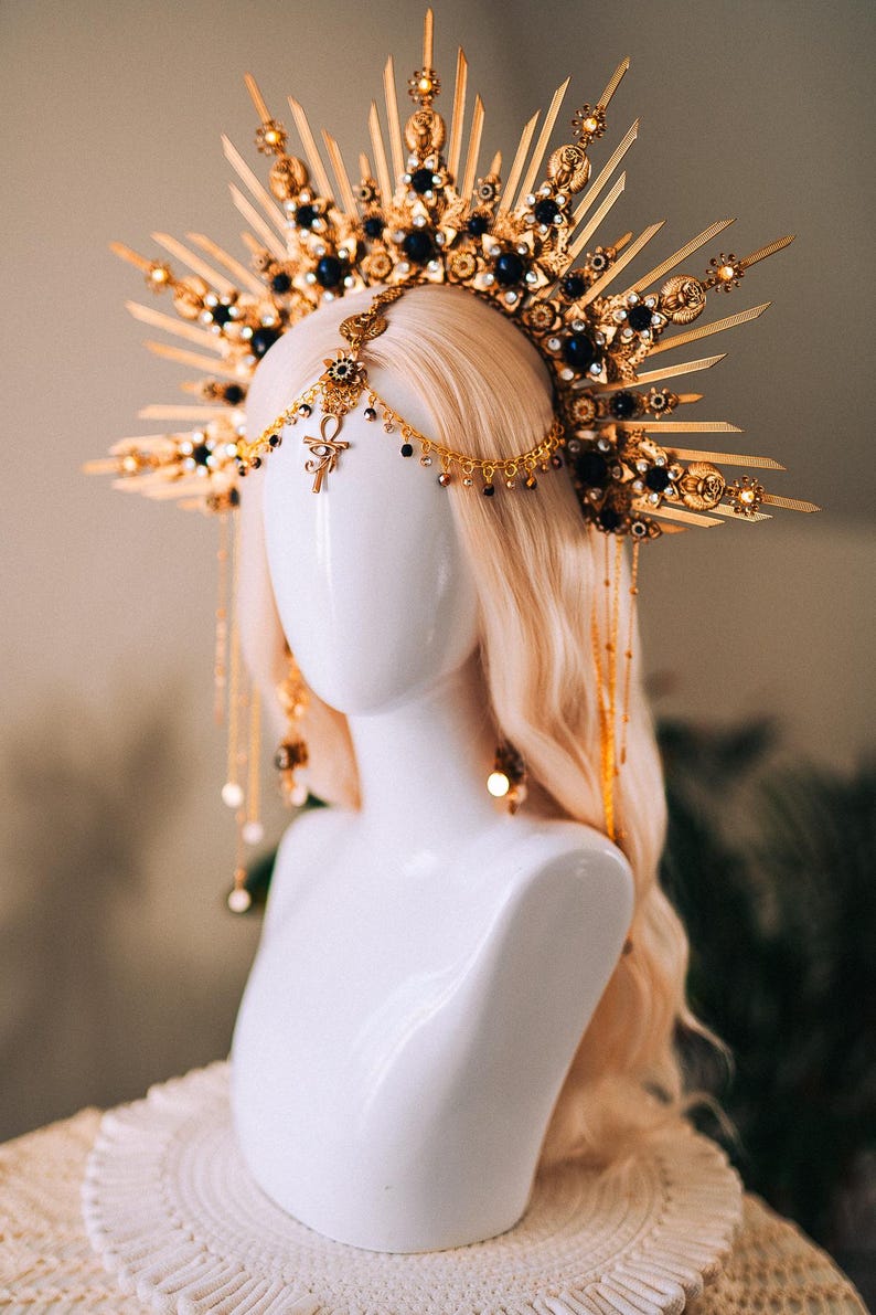 May include: A gold-toned headpiece with a sunburst design, adorned with dark blue gemstones and dangling chains. The headpiece features an Ankh symbol and is displayed on a white mannequin head with blonde hair, showcasing a celestial or fantasy theme.