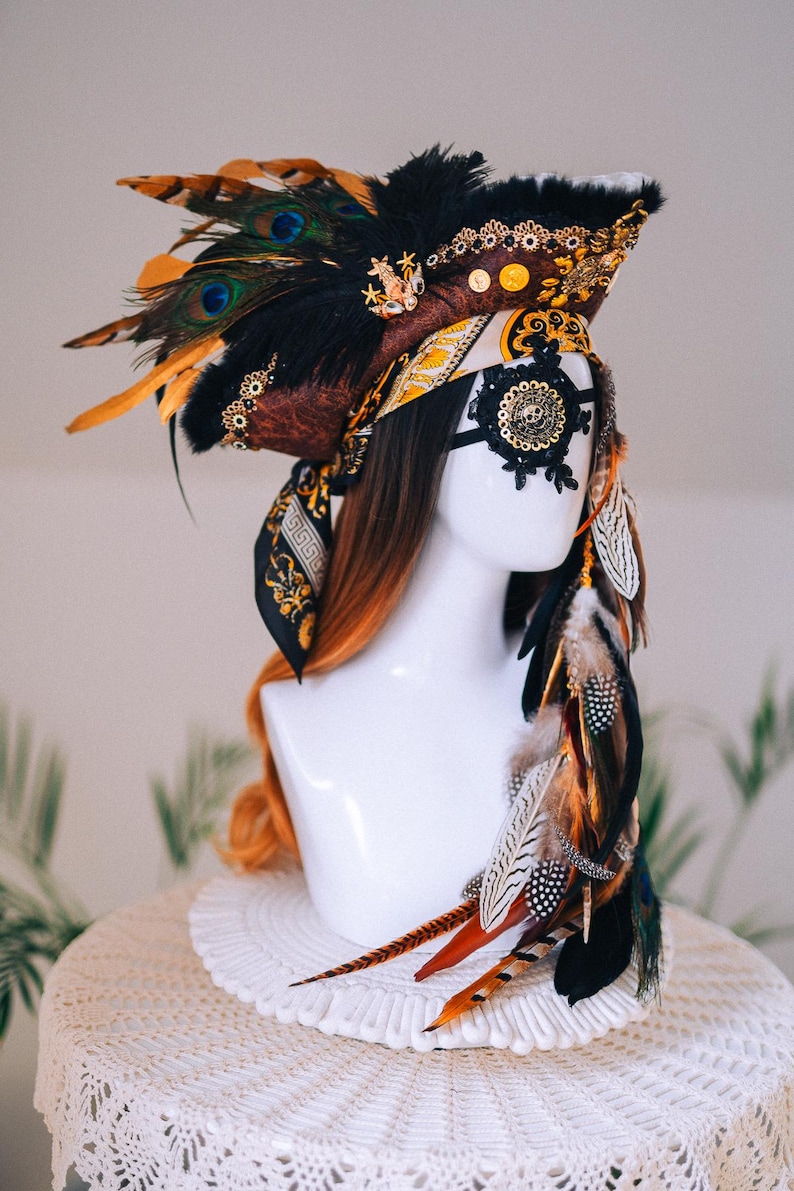Puede incluir: Tocado decorativo con plumas de pavo real, detalles dorados y una m&aacute;scara de encaje negro. El tocado presenta una combinaci&oacute;n de colores negro, marr&oacute;n y dorado. Plumas adicionales caen por el costado, creando un aspecto dram&aacute;tico y ornamentado.