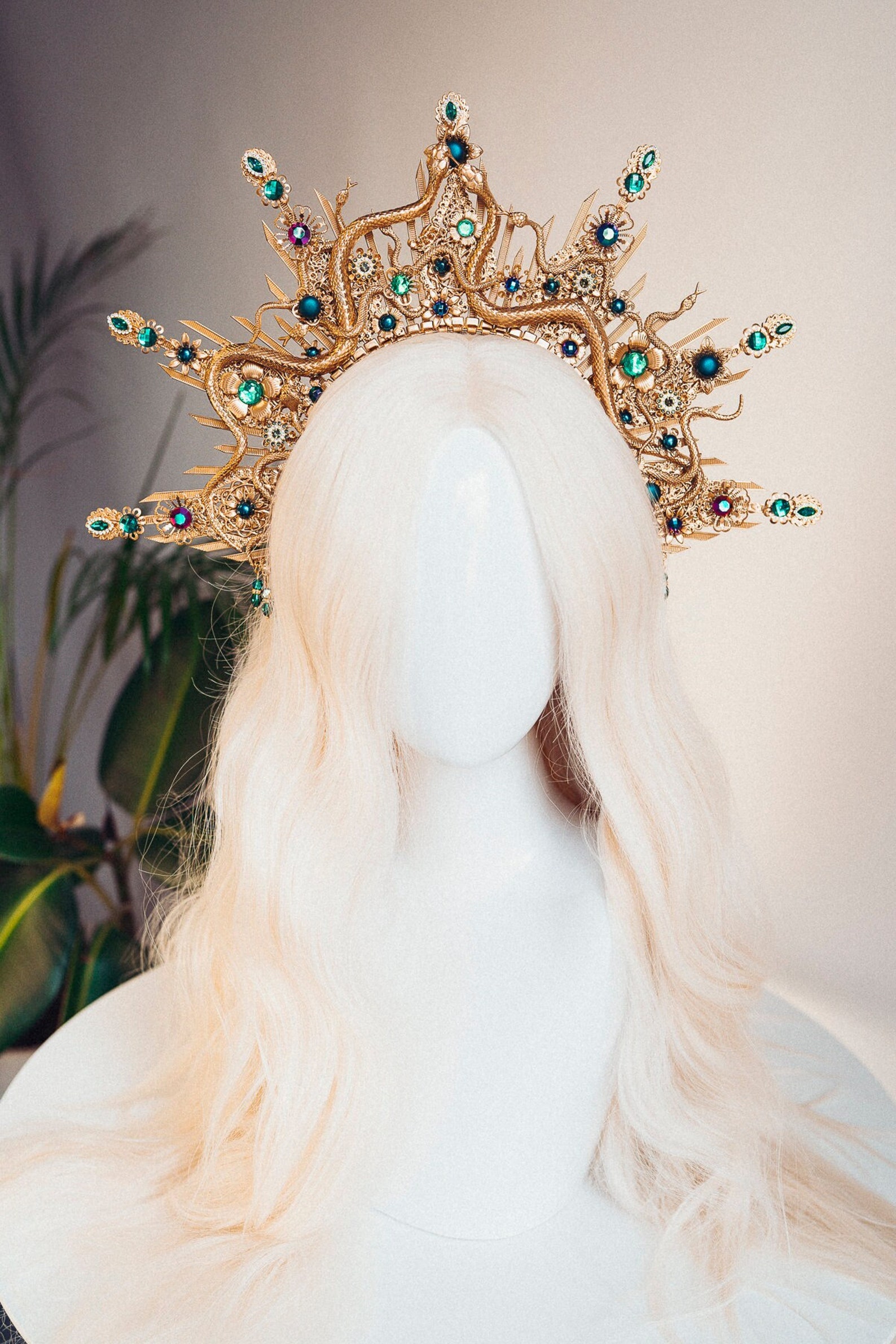 White wig on a mannequin head. A crown made out of gold snakes and green jewels covers it.