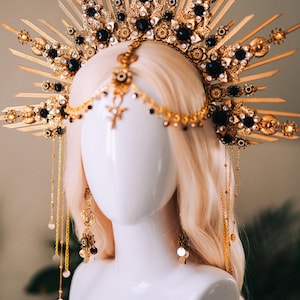May include: A gold-colored headpiece with intricate detailing, featuring dark blue gemstones and ornate embellishments. The headpiece is displayed on a mannequin head with blonde hair, showcasing its design and craftsmanship. The piece includes dangling chains and earrings.