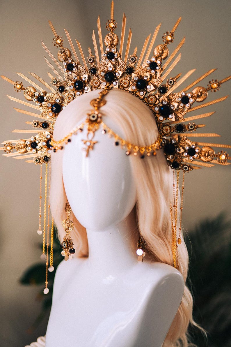 May include: A gold-colored headpiece with intricate detailing, featuring dark blue gemstones and ornate embellishments. The headpiece is displayed on a mannequin head with blonde hair, showcasing its design and craftsmanship. The piece includes dangling chains and earrings.