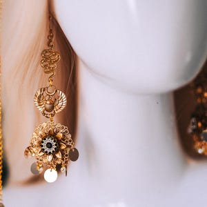 May include: Gold-toned statement earrings with an Egyptian theme. The earrings feature intricate designs, including a scarab beetle, floral elements, and dangling circular charms. The earrings are displayed on a white mannequin head.