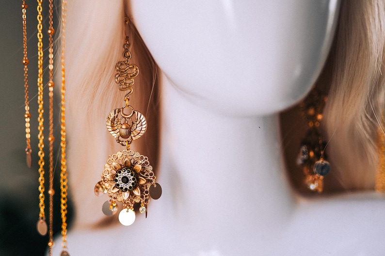 May include: Gold-toned statement earrings with an Egyptian theme. The earrings feature intricate designs, including a scarab beetle, floral elements, and dangling circular charms. The earrings are displayed on a white mannequin head.