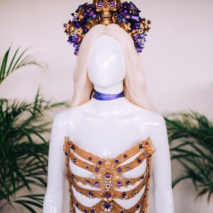 May include: A gold and purple jeweled bodice with a skull and flower crown. The bodice is made of lace and features a ribcage design. The crown is adorned with purple flowers and a gold skull with jeweled eyes.