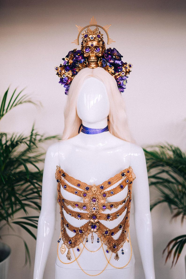 May include: A gold and purple jeweled bodice with a skull and flower crown. The bodice is made of lace and features a ribcage design. The crown is adorned with purple flowers and a gold skull with jeweled eyes.