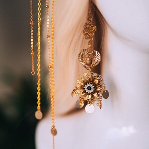 May include: Ornate gold-toned earrings with intricate designs and dangling chains. The earrings feature a floral element with a central black gem and circular accents. The design includes a scarab beetle motif and delicate chain details.
