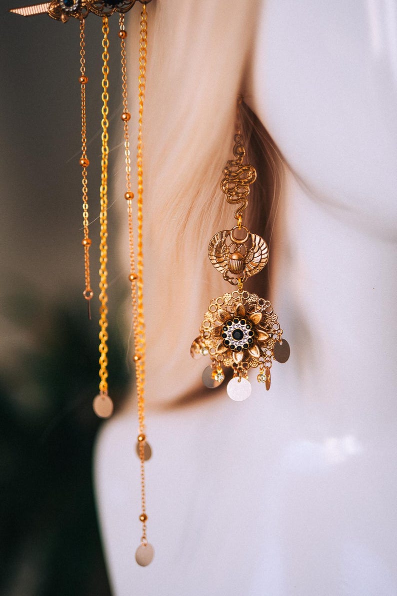 May include: Ornate gold-toned earrings with intricate designs and dangling chains. The earrings feature a floral element with a central black gem and circular accents. The design includes a scarab beetle motif and delicate chain details.