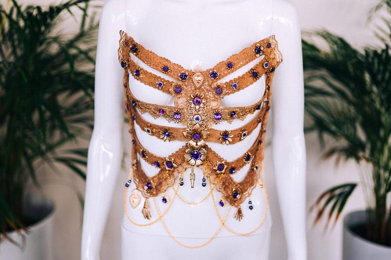 May include: A gold-toned, beaded, and lace body chain with purple gemstones. The chain is layered and has a delicate, feminine design.