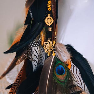 Puede incluir: Un accesorio decorativo para el cabello con una mezcla de plumas en negro, marr&oacute;n, naranja y blanco. Se adjuntan amuletos dorados, incluyendo una br&uacute;jula y un dise&ntilde;o floral.