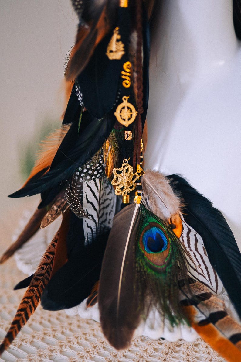 Puede incluir: Un accesorio decorativo para el cabello con una mezcla de plumas en negro, marr&oacute;n, naranja y blanco. Se adjuntan amuletos dorados, incluyendo una br&uacute;jula y un dise&ntilde;o floral.