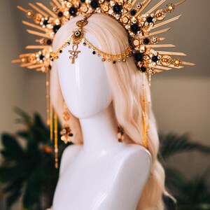 May include: A gold-toned headpiece with a sunburst design, adorned with black and gold accents. The headpiece features dangling chains and an Ankh symbol, displayed on a white mannequin head with blonde hair. A unique accessory.