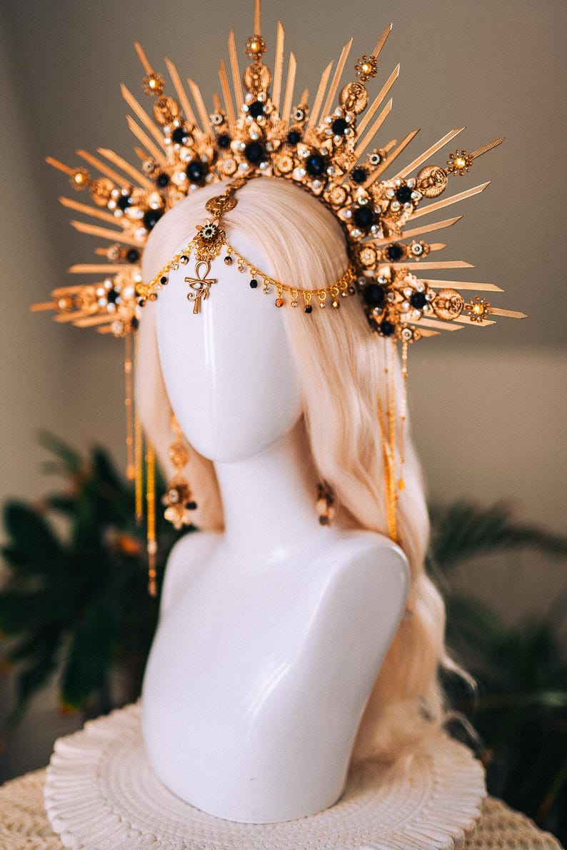 May include: A gold-toned headpiece with a sunburst design, adorned with black and gold accents. The headpiece features dangling chains and an Ankh symbol, displayed on a white mannequin head with blonde hair. A unique accessory.