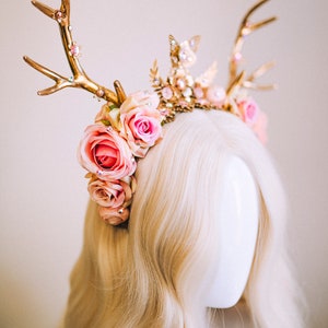 Puede incluir: Una diadema de asta de ciervo dorada con rosas rosas y detalles de perlas. La diadema se lleva en una peluca rubia.