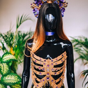 May include: A gold and purple skeleton-shaped bodice with a skull and flower crown. The bodice is made of lace and features a variety of purple gemstones. The crown is adorned with purple flowers and a gold skull with purple gemstones for eyes.