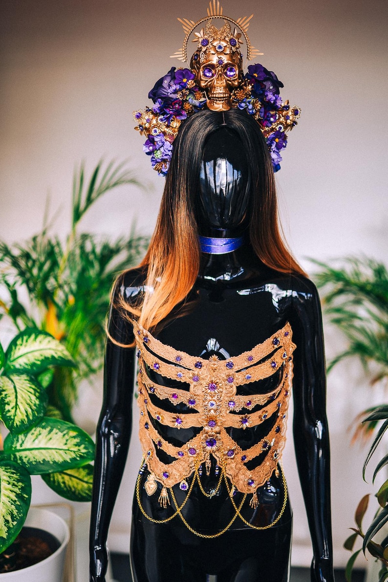 May include: A gold and purple skeleton-shaped bodice with a skull and flower crown. The bodice is made of lace and features a variety of purple gemstones. The crown is adorned with purple flowers and a gold skull with purple gemstones for eyes.