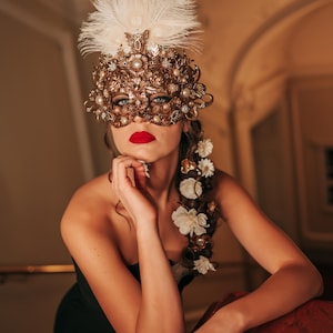 K&ouml;nnte beinhalten: Eine Frau tr&auml;gt eine goldene und wei&szlig;e Maskenballmaske mit Federn und einem Blumenhaarschmuck. Sie tr&auml;gt ein schwarzes tr&auml;gerloses Kleid und roten Lippenstift.