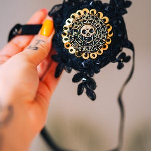 Puede incluir: M&aacute;scara de encaje negro con un dise&ntilde;o de calavera y sol dorado. La m&aacute;scara presenta un emblema circular central con una calavera y patrones intrincados, rodeado de lentejuelas doradas. La m&aacute;scara est&aacute; adornada con encaje negro y elementos decorativos.
