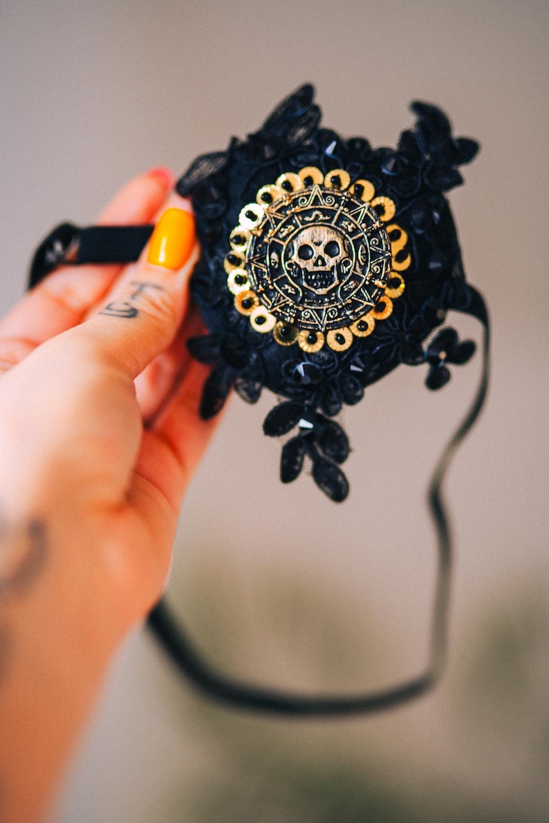 Puede incluir: M&aacute;scara de encaje negro con un dise&ntilde;o de calavera y sol dorado. La m&aacute;scara presenta un emblema circular central con una calavera y patrones intrincados, rodeado de lentejuelas doradas. La m&aacute;scara est&aacute; adornada con encaje negro y elementos decorativos.