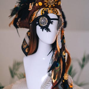 Puede incluir: Un tocado decorativo con tem&aacute;tica pirata. El tocado presenta un sombrero marr&oacute;n y negro con adornos dorados, un parche en el ojo negro y una variedad de plumas en tonos marrones, naranjas y verdes. El tocado se exhibe en una cabeza de maniqu&iacute; blanca.