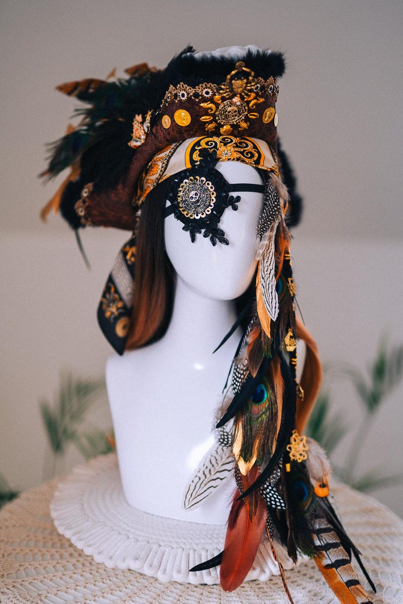 Puede incluir: Un tocado decorativo con tem&aacute;tica pirata. El tocado presenta un sombrero marr&oacute;n y negro con adornos dorados, un parche en el ojo negro y una variedad de plumas en tonos marrones, naranjas y verdes. El tocado se exhibe en una cabeza de maniqu&iacute; blanca.