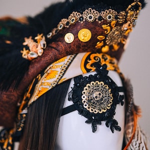 Puede incluir: Un sombrero de pirata decorativo con una corona marr&oacute;n con aspecto de cuero, adornado con monedas de oro, joyas y plumas negras. Se adjunta un parche en el ojo de encaje negro con un dise&ntilde;o de calavera dorada. El sombrero es parte de un disfraz.