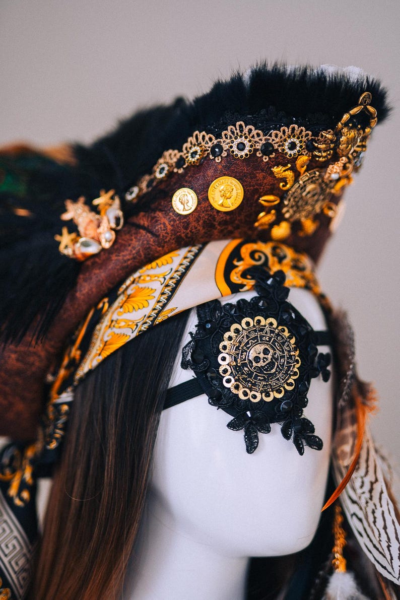 Puede incluir: Un sombrero de pirata decorativo con una corona marr&oacute;n con aspecto de cuero, adornado con monedas de oro, joyas y plumas negras. Se adjunta un parche en el ojo de encaje negro con un dise&ntilde;o de calavera dorada. El sombrero es parte de un disfraz.