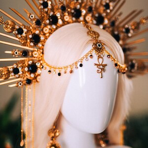 May include: A gold-colored headpiece with black accents and a sunburst design. The headpiece features dangling chains and an Ankh symbol. The jewelry is displayed on a white mannequin head with blonde hair.
