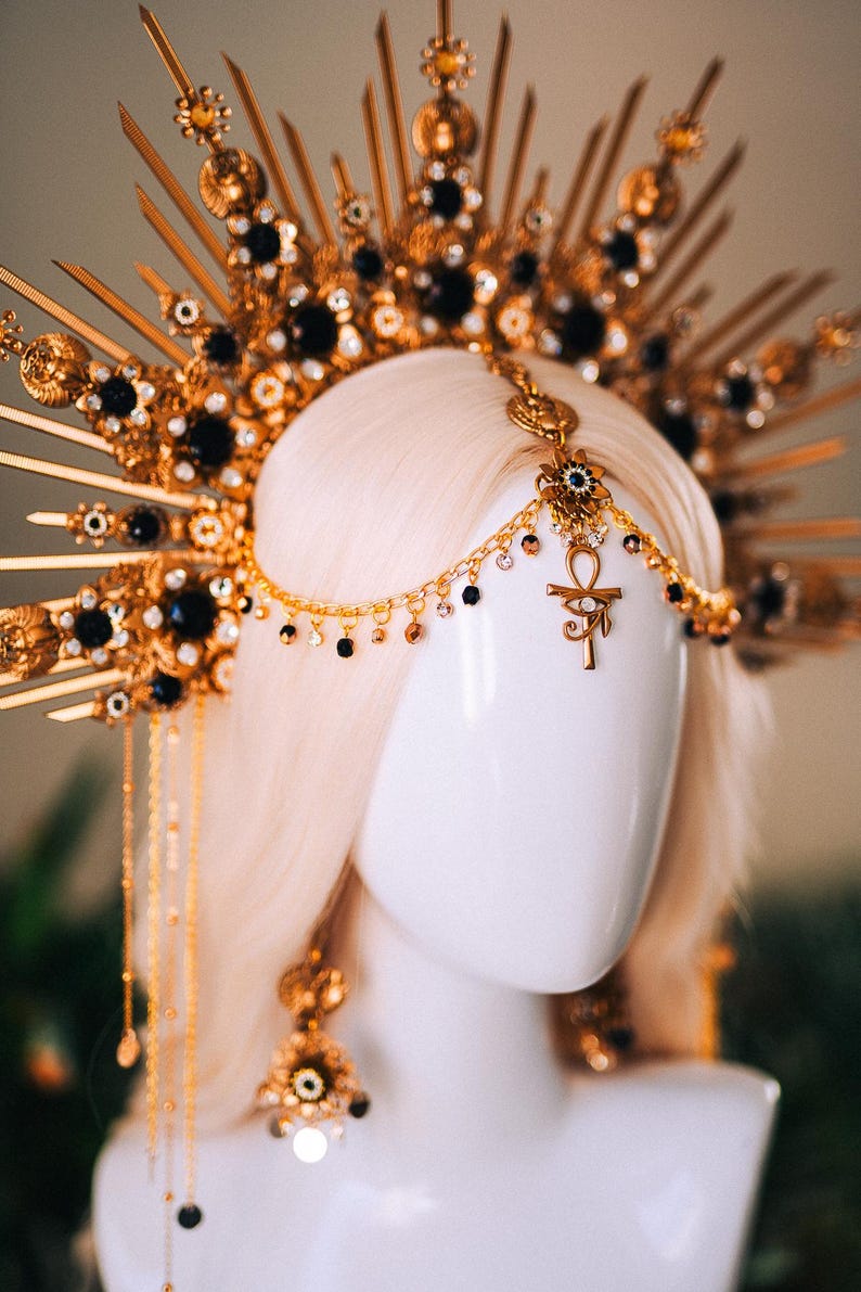 May include: A gold-colored headpiece with black accents and a sunburst design. The headpiece features dangling chains and an Ankh symbol. The jewelry is displayed on a white mannequin head with blonde hair.