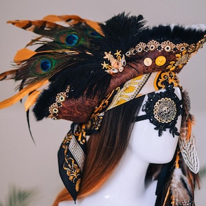 Puede incluir: Un tocado decorativo con tem&aacute;tica pirata, con un sombrero marr&oacute;n adornado con plumas negras, plumas de pavo real y adornos dorados. Una m&aacute;scara para los ojos negra con un dise&ntilde;o de calavera y una bufanda estampada completan el look.