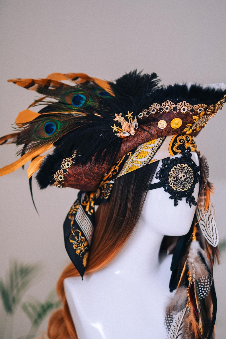 Puede incluir: Un tocado decorativo con tem&aacute;tica pirata, con un sombrero marr&oacute;n adornado con plumas negras, plumas de pavo real y adornos dorados. Una m&aacute;scara para los ojos negra con un dise&ntilde;o de calavera y una bufanda estampada completan el look.