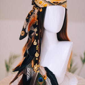 Puede incluir: Un tocado de estilo bohemio con un pa&ntilde;uelo estampado en negro, dorado y crema. Adornado con plumas marrones, negras y naranjas, y abalorios dorados. Una pluma de pavo real a&ntilde;ade un toque de color. Estilo bohemio.