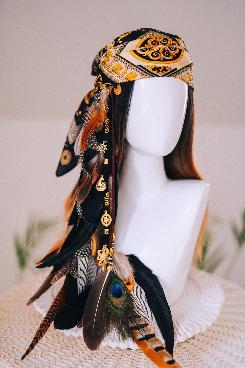 Puede incluir: Un tocado de estilo bohemio con un pa&ntilde;uelo estampado en negro, dorado y crema. Adornado con plumas marrones, negras y naranjas, y abalorios dorados. Una pluma de pavo real a&ntilde;ade un toque de color. Estilo bohemio.