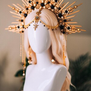 May include: A gold and black headpiece with a sunburst design, adorned with black gemstones and dangling gold chains. The headpiece is displayed on a white mannequin head with long, wavy blonde hair. The headpiece features an Ankh symbol.