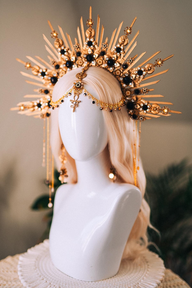 May include: A gold and black headpiece with a sunburst design, adorned with black gemstones and dangling gold chains. The headpiece is displayed on a white mannequin head with long, wavy blonde hair. The headpiece features an Ankh symbol.