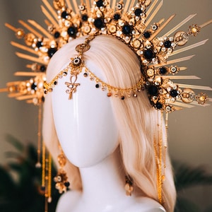 May include: A gold-toned headpiece with a sunburst design, adorned with black gemstones and intricate detailing. The headpiece features dangling chains and an Ankh symbol, creating an elegant and celestial aesthetic. The headpiece is displayed on a mannequin head with blonde hair.