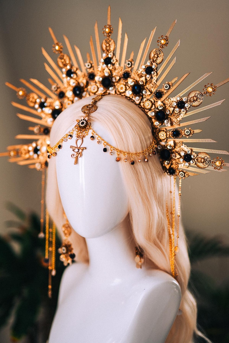 May include: A gold-toned headpiece with a sunburst design, adorned with black gemstones and intricate detailing. The headpiece features dangling chains and an Ankh symbol, creating an elegant and celestial aesthetic. The headpiece is displayed on a mannequin head with blonde hair.